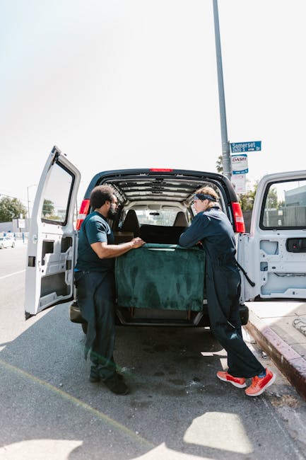 Two movers from Man with Van New Eltham are engaged in a home relocation process, loading a large, wrapped piece of furniture or appliance into the open rear of a white cargo van parked on a paved street. The interior of the van shows a spacious cargo area with a black rubber mat and some packaging materials. One mover is holding the furniture with both hands, while the other is guiding it into the vehicle. They are dressed in dark uniforms with safety glasses, and the moving process is taking place on a sunny day near urban surroundings, including a street sign and a sidewalk curb. This scene depicts furniture transport and packing activities involved in a professional removals service at the doorstep during a same-day move for a property in New Eltham, consistent with the offerings of Man with Van New Eltham.