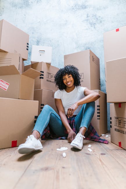 A young woman with curly black hair and a bright smile is seated on a wooden floor surrounded by large cardboard boxes used for house removals and furniture transport. She is wearing a white t-shirt, light blue jeans, and white sneakers, with a plaid shirt tied around her waist. The boxes vary in size and are stacked behind her against a light blue textured wall, some with red and blue markings indicating fragile contents. Small pieces of packing material are scattered on the floor near her feet. In this interior setting, the image captures a moment during packing or early stages of a home relocation, with the woman in a relaxed pose amidst the moving supplies. Occasionally, Man with Van New Eltham offers removals and packing services, including furniture transport and loading, as part of their house removal process, reflected in the visual scene.