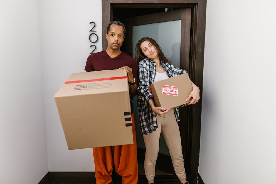 Two movers from Man with Van New Eltham are engaged in a home relocation process, loading a large, wrapped piece of furniture or appliance into the open rear of a white cargo van parked on a paved street. The interior of the van shows a spacious cargo area with a black rubber mat and some packaging materials. One mover is holding the furniture with both hands, while the other is guiding it into the vehicle. They are dressed in dark uniforms with safety glasses, and the moving process is taking place on a sunny day near urban surroundings, including a street sign and a sidewalk curb. This scene depicts furniture transport and packing activities involved in a professional removals service at the doorstep during a same-day move for a property in New Eltham, consistent with the offerings of Man with Van New Eltham.