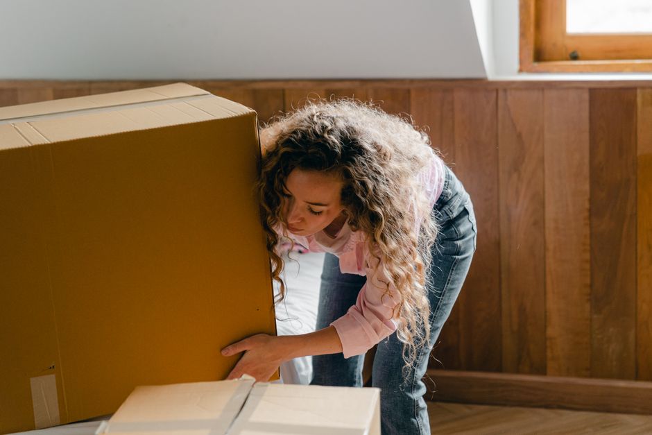 A woman with curly hair, wearing a light pink shirt and blue jeans, is in a room with wooden paneling on the walls and a window with natural light. She is bending over, actively lifting or adjusting a large cardboard box, which is part of a home relocation or furniture transport process. The box appears sturdy and is placed on a floor that may be wooden or laminate. Nearby, in the foreground, there is a smaller open cardboard box, possibly containing packing materials. The scene suggests the interior of a house in the midst of packing or moving activities, with the woman engaged in preparing items for a house removals service. The image captures the physical effort involved in packing and the careful handling of household items, aligning with professional removal activities provided by companies like Man with Van New Eltham.