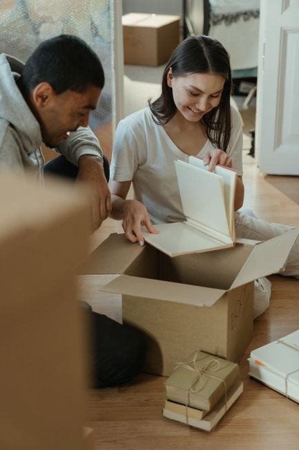 A young woman and a man are inside a home during a house relocation, sitting on a wooden floor surrounded by open cardboard boxes filled with books and packaging materials. The woman is holding a whit