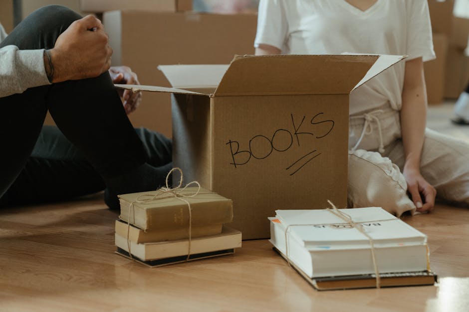 A young woman and a man are inside a home during a house relocation, sitting on a wooden floor surrounded by open cardboard boxes filled with books and packaging materials. The woman is holding a whit