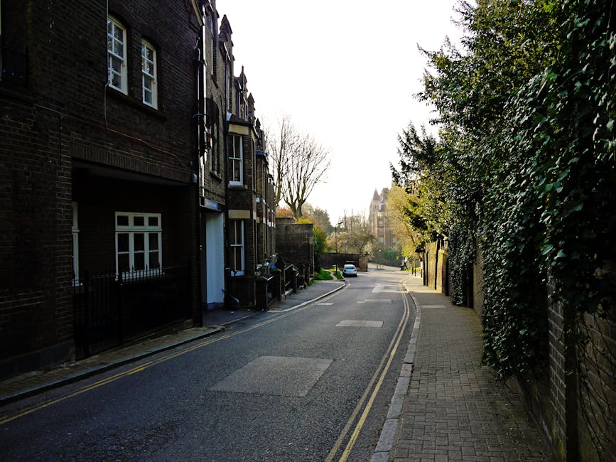 A narrow residential road lined with tall, leafy trees on both sides, creating a canopy overhead. The pavement is flanked by a metal fence on the left, behind which dense shrubbery and greenery are vi