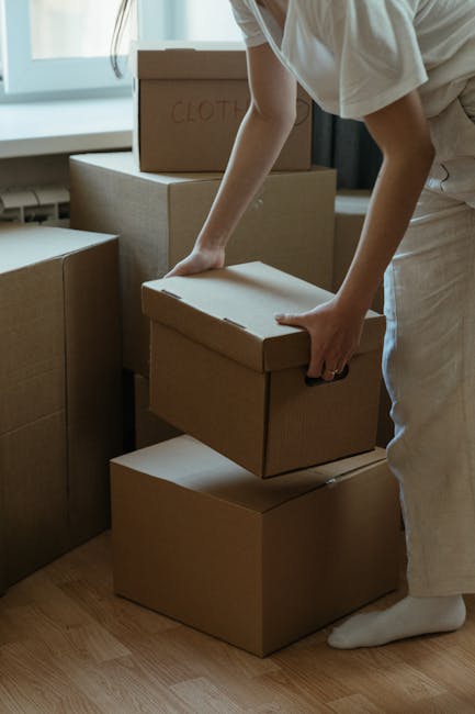 A person is seen in an indoor setting, using their hands to lift a cardboard box that is part of a stack of moving boxes. The boxes are of various sizes, some labeled with markings such as 'clothes,' 