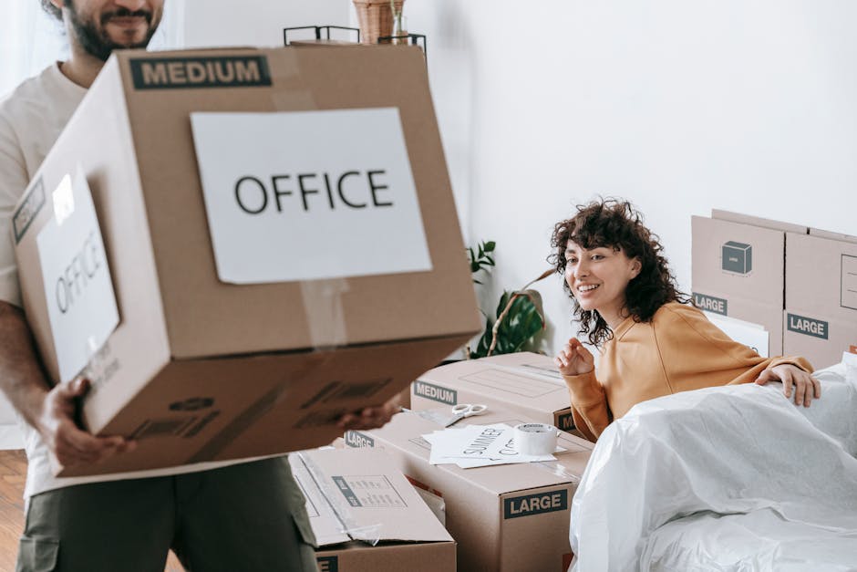 A person is seen in an indoor setting, using their hands to lift a cardboard box that is part of a stack of moving boxes. The boxes are of various sizes, some labeled with markings such as 'clothes,' 